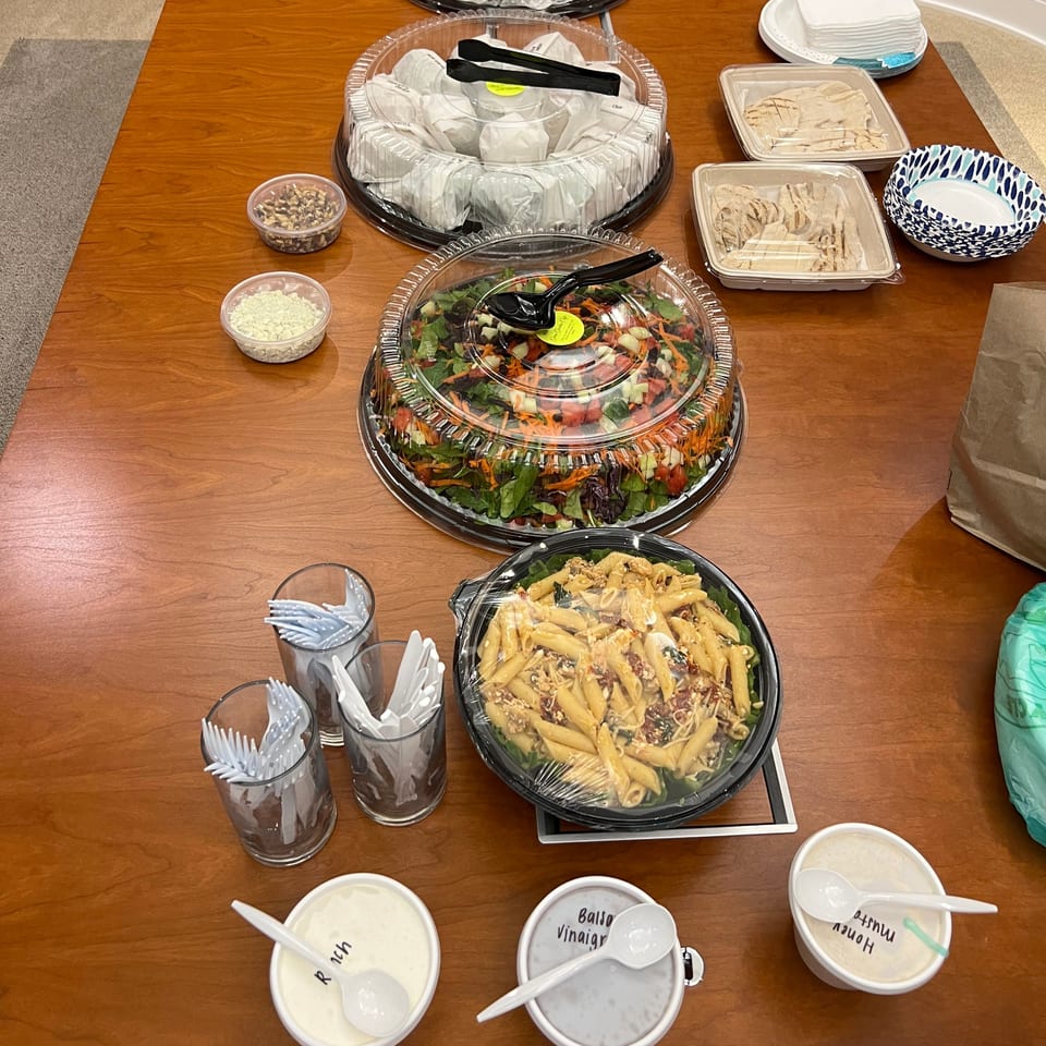 A catering spread on a wooden table with salads, pasta, sandwiches, and dressings in disposable containers.