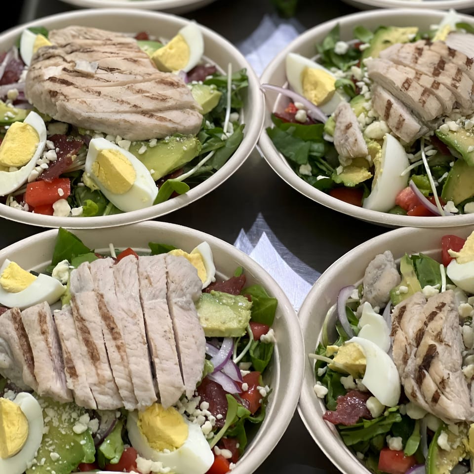 Four grilled chicken salads with avocado, hard-boiled eggs, and tomatoes in white bowls.