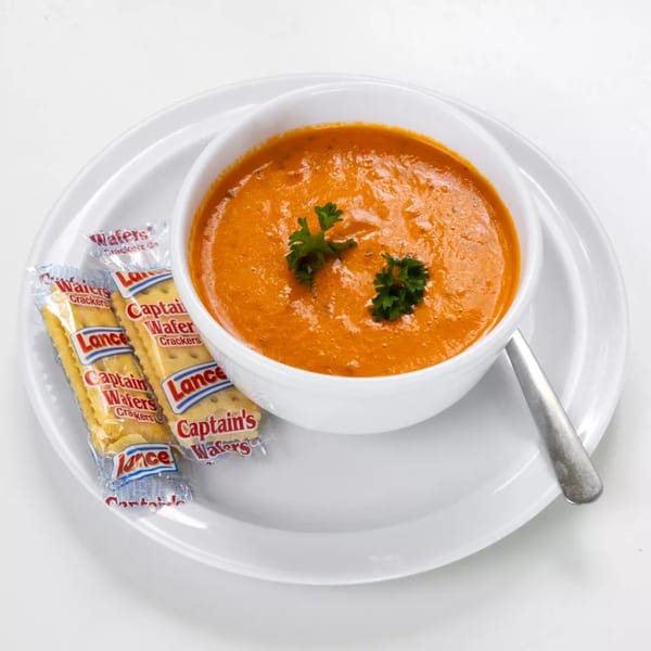 A white bowl of creamy tomato soup garnished with parsley, served with two packets of Lance crackers.