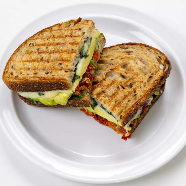 Grilled sandwich with avocado, cheese, spinach, and sun-dried tomatoes on seeded bread, cut in half.