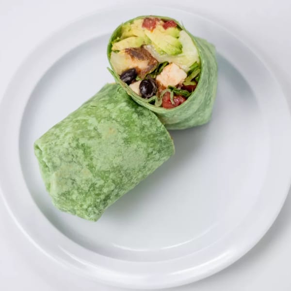 A green tortilla wrap, cut in half, filled with chicken, avocado, black olives, and tomatoes.