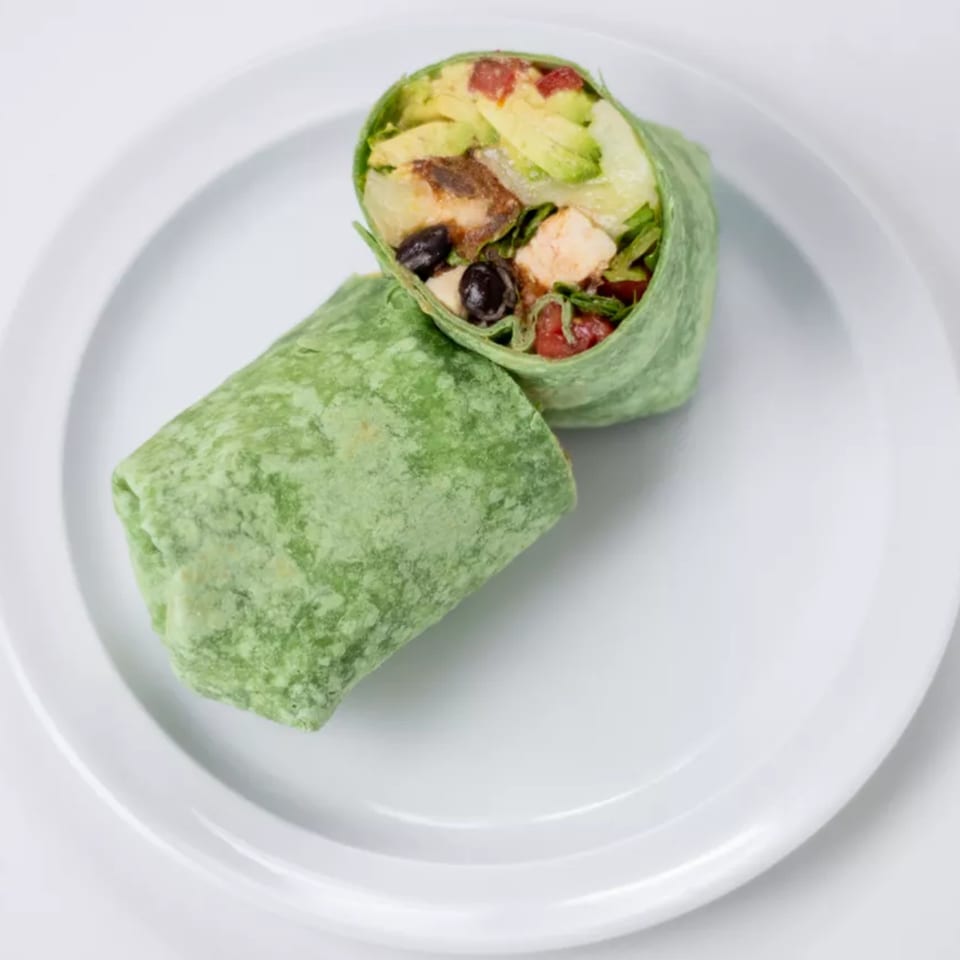 A green tortilla wrap, cut in half, filled with chicken, avocado, black olives, and tomatoes.